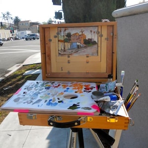 May include: A wooden artist's field easel with a painting in progress, a palette of mixed paints, and various brushes. The painting depicts a street scene with buildings and palm trees. The easel is set up outdoors.