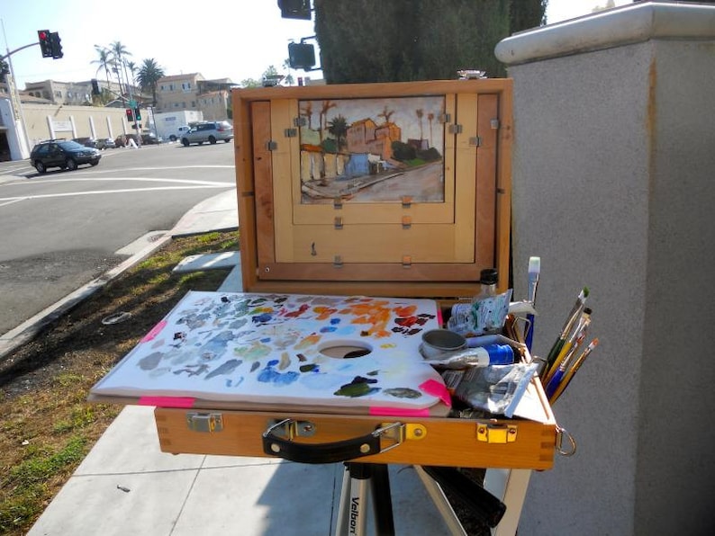 May include: A wooden artist's field easel with a painting in progress, a palette of mixed paints, and various brushes. The painting depicts a street scene with buildings and palm trees. The easel is set up outdoors.