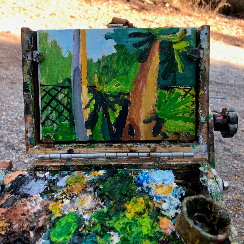 Puede incluir: Una caja de pintura de artista de madera abierta con una pintura de paisaje de &aacute;rboles y un cielo azul. La paleta est&aacute; llena de varios colores de pintura. La pintura est&aacute; en un marco de madera.