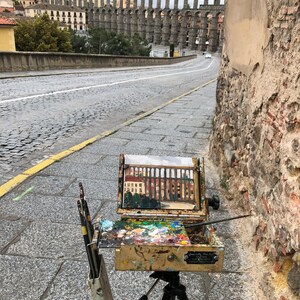 May include: A wooden artist's easel with a canvas depicting an aqueduct, set up on a cobblestone street with a view of the aqueduct in the distance.