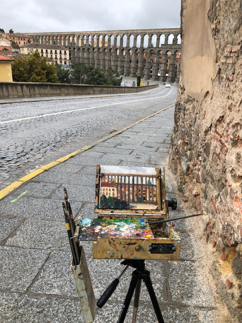 May include: A wooden artist's easel with a canvas depicting an aqueduct, set up on a cobblestone street with a view of the aqueduct in the distance.