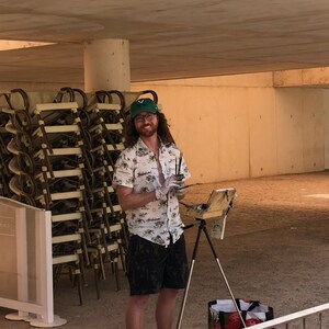 May include: A man wearing a green baseball cap and a floral shirt is painting outdoors. He is standing in front of a stack of chairs and using a paintbrush and palette. He is standing on a concrete surface with a tripod easel and a bag of supplies.
