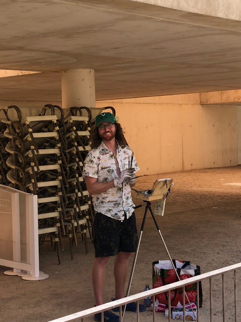 May include: A man wearing a green baseball cap and a floral shirt is painting outdoors. He is standing in front of a stack of chairs and using a paintbrush and palette. He is standing on a concrete surface with a tripod easel and a bag of supplies.