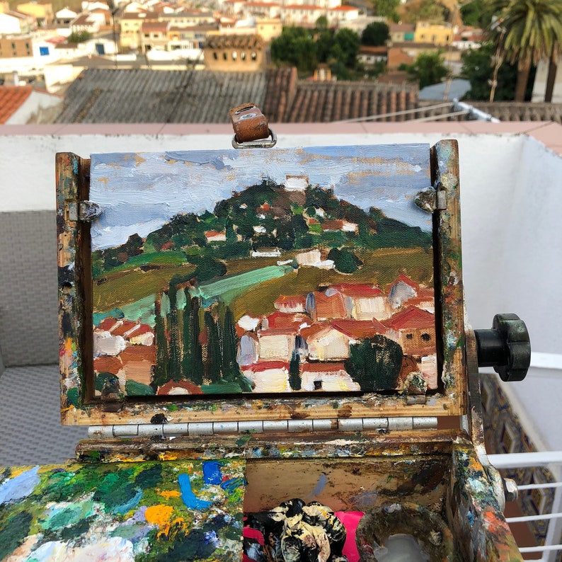 May include: A small, wooden easel with a hinged lid, open to reveal a painting of a hillside village. The painting is done in oil paints and features a variety of colors, including green, brown, and white. The easel is sitting on a surface that appears to be a rooftop.