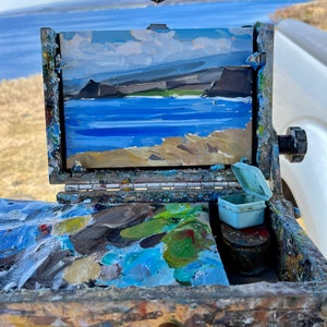 May include: An open wooden artist's paint box with a small landscape painting of a coastal scene. The painting depicts a blue ocean, tan cliffs, and a cloudy sky. The box contains a palette of mixed paint colors and small containers.