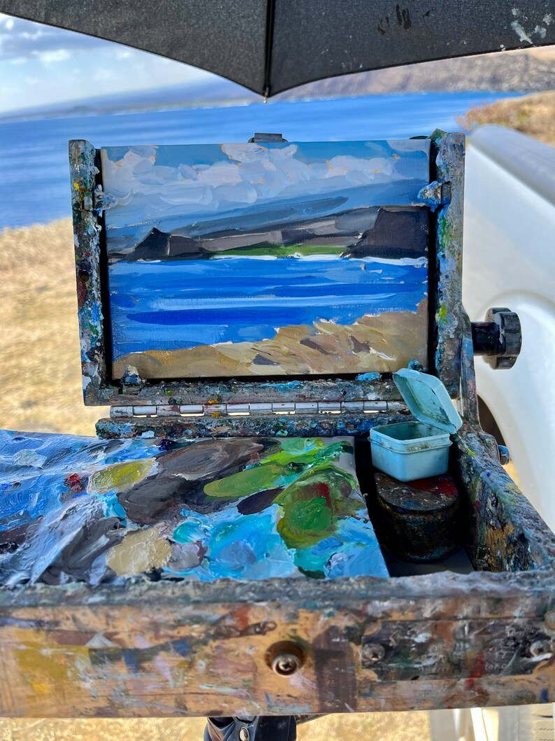 May include: An open wooden artist's paint box with a small landscape painting of a coastal scene. The painting depicts a blue ocean, tan cliffs, and a cloudy sky. The box contains a palette of mixed paint colors and small containers.