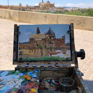 May include: An open wooden artist's box with a small painting of a building with a dome. The painting is in a wooden frame. The box contains a palette of mixed paint colors, brushes, and other art supplies. The background shows a similar building.