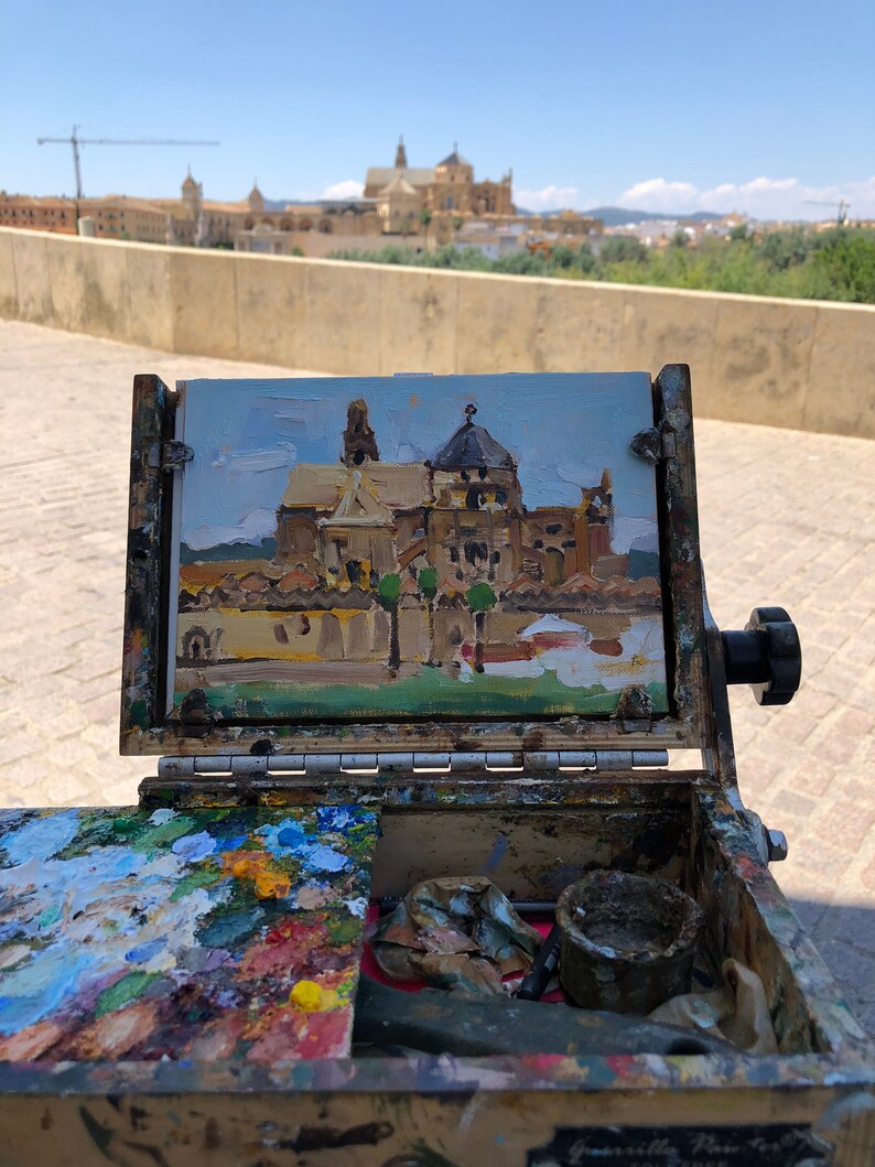 May include: An open wooden artist's box with a small painting of a building with a dome. The painting is in a wooden frame. The box contains a palette of mixed paint colors, brushes, and other art supplies. The background shows a similar building.