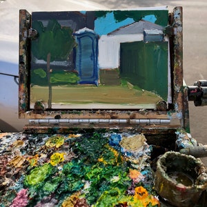 Puede incluir: Una pintura al &oacute;leo de un ba&ntilde;o port&aacute;til azul y un edificio blanco con un seto verde. La pintura est&aacute; sobre un caballete con una paleta de colores de pintura mezclados, incluyendo verde, amarillo y rosa.