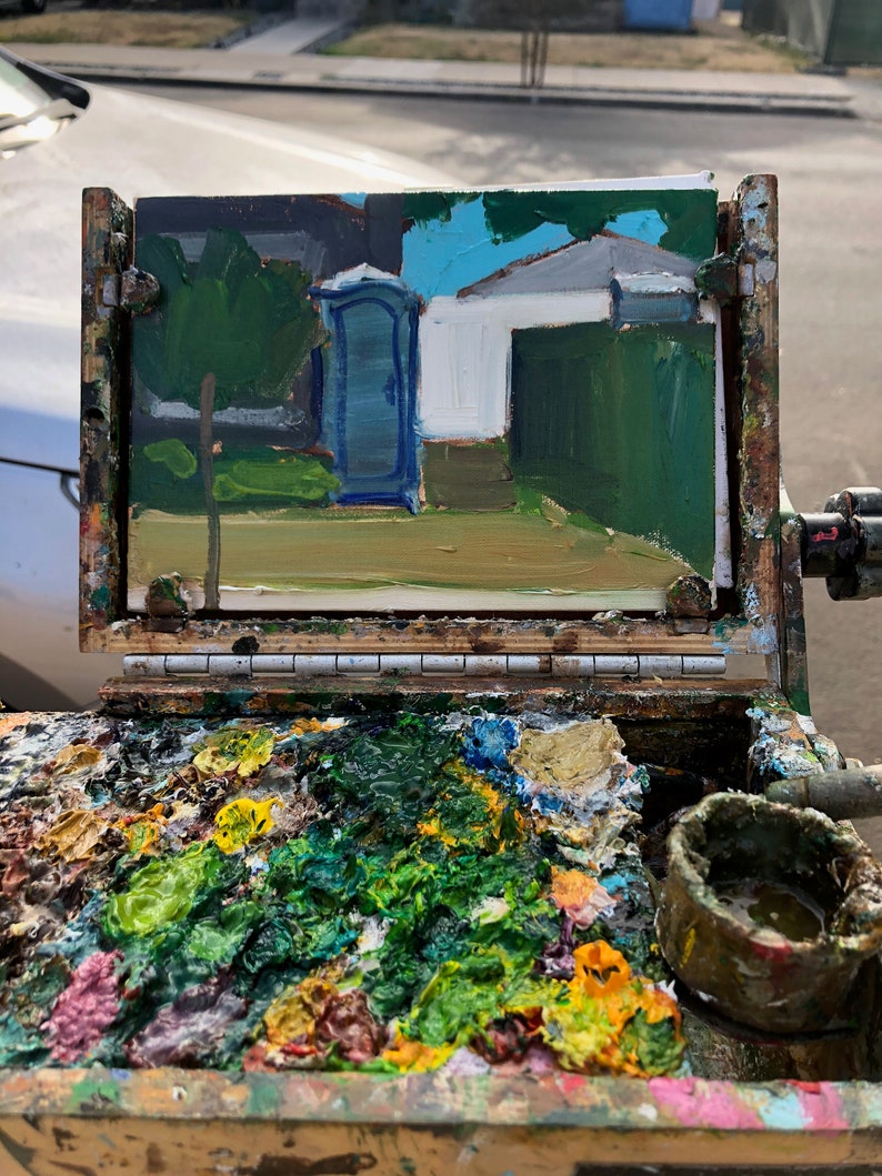Puede incluir: Una pintura al &oacute;leo de un ba&ntilde;o port&aacute;til azul y un edificio blanco con un seto verde. La pintura est&aacute; sobre un caballete con una paleta de colores de pintura mezclados, incluyendo verde, amarillo y rosa.