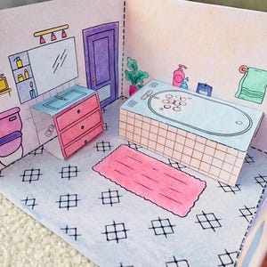 May include: A papercraft bathroom scene with a pink rug, a light blue bathtub, and a pink sink cabinet. The walls are light pink with black line art details. The floor has a black and white pattern.