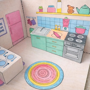 May include: A colorful papercraft kitchen scene with a pink refrigerator, teal cabinets, and a stove. The kitchen includes a sink, blender, and a rolling pin. A round rug with pink, yellow, and blue spirals sits on the floor.