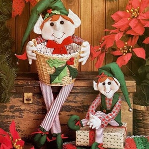 May include: Two plush Christmas elves, one holding a wicker basket with holly and the other sitting on a wrapped gift. Both elves are wearing red and green plaid outfits and have green hats with white trim.