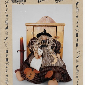 Puede incluir: Un patrón de muñeca bruja llamado "Chelsea" de "Katie's Homespun Stitches". La muñeca mide 45,7 cm de alto y está sentada frente a un armario de madera. La muñeca lleva un vestido negro y marrón con un delantal blanco y un sombrero de bruja negro.
