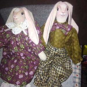 May include: Two handmade fabric bunny dolls dressed in vintage-style clothing. The doll on the left is wearing a purple floral dress with a white lace collar. The doll on the right is wearing a green and brown plaid shirt with a brown bow tie and matching plaid pants.