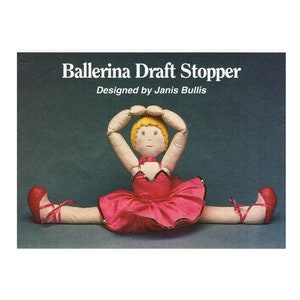 May include: A ballerina doll draft stopper with a pink tutu and red shoes. The doll has blonde hair and is holding her arms above her head. The text "Ballerina Draft Stopper" and "Designed by Janis Bullis" is printed on the image.