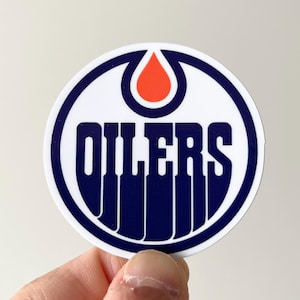 Op de afbeelding: Een ronde sticker met het logo van de Edmonton Oilers, bestaande uit een donkerblauwe oliedruppel bovenaan, een witte achtergrond en het woord OILERS in dikke, donkerblauwe hoofdletters met verticale lijnen die naar beneden lopen.