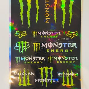Puede incluir: Una hoja de pegatinas holográficas con el logotipo de Monster Energy en varios tamaños y orientaciones, incluyendo la icónica garra, la palabra Monster y el logo de Fox Racing, con el texto ZC-2Y-01 impreso sobre el fondo negro.