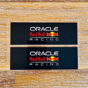 Może przedstawiać: Dwie prostokątne czarne naklejki z logo Oracle Red Bull Racing w kolorze białym, czerwonym i żółtym. Napis Oracle widnieje w kolorze białym, za nim znajduje się logo Red Bull z dwoma czerwonymi bykami i żółtym słońcem oraz słowo Racing.