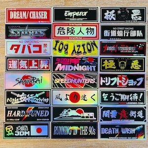 May include: A collection of rectangular vinyl stickers featuring various Japanese-inspired automotive and street culture graphics, text, and logos in diverse colours, fonts, and holographic finishes arranged in a grid on a light wooden surface.