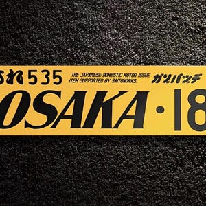 May include: A rectangular yellow sticker featuring black text that reads OSAKA 18, 535, and THE JAPANESE DOMESTIC MOTOR ISSUE ITEM SUPPORTED BY SAITOWORKS, along with Japanese characters and a small Original Goods logo in the top right corner.