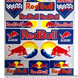 Peut inclure: Une feuille d'autocollants Red Bull avec des taureaux rouges, des soleils jaunes et des fonds bleus, incluant le texte Red Bull et The Red Bulletin. Une règle en plastique en bas indique une largeur de feuille d'environ 24 cm.