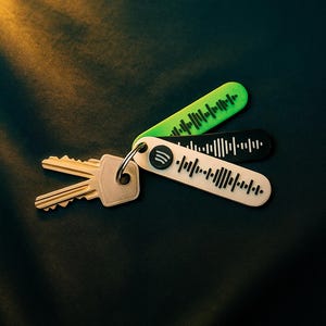 Könnte beinhalten: Ein Schlüsselbund mit einem silbernen Schlüsselring und drei Schlüsselanhängern im Spotify-Design. Die Schlüsselanhänger sind grün, schwarz und weiß, jeder mit einem einzigartigen Schallwellenmuster. Der Hintergrund ist eine dunkle, strukturierte Oberfläche.