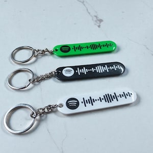 Könnte beinhalten: Drei Schlüsselanhänger mit Spotify-Code-Designs. Einer ist grün, einer schwarz und einer weiß. Jeder Schlüsselanhänger hat einen silberfarbenen Ring und eine Kette. Die Schlüsselanhänger sind länglich und haben ein strichcodeähnliches Design.
