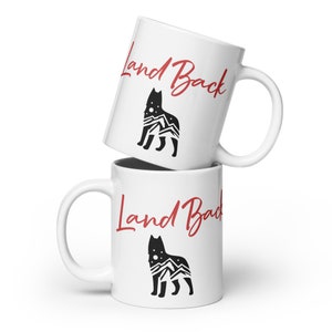 May include: Two white ceramic mugs stacked on top of each other. The top mug has the text "Land Back" in red cursive script with a black silhouette of a wolf with mountains and stars inside. The bottom mug has the same design but the text is in red cursive script and the wolf is facing the opposite direction.