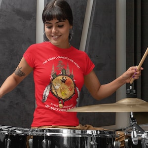 May include: A woman wearing a red t-shirt with the text "The drum connects our heart to the heartbeat of Mother Earth" and a graphic of a drum with feathers and handprints. She is playing drums with two drumsticks.