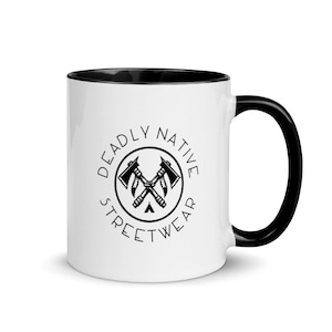 Könnte beinhalten: Weißer Keramikbecher mit schwarzem Innenraum und Henkel. Der Becher zeigt den Text "DEADLY NATIVE STREETWEAR" in einem kreisförmigen Design mit gekreuzten Äxten in der Mitte. Das Design ist in Schwarz.