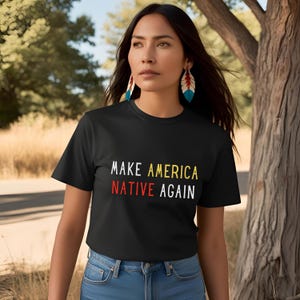 May include: A black t-shirt with the text "MAKE AMERICA NATIVE AGAIN" in white, yellow, and red lettering. The shirt is being worn by a person with long dark hair and feather earrings.