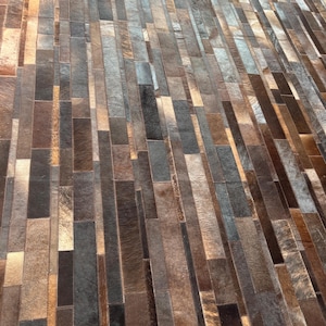 Puede incluir: Una alfombra rectangular con un diseño de patchwork. La alfombra está hecha de piezas rectangulares de piel en tonos marrones, grises y beige. Las piezas están dispuestas en un patrón lineal, creando una superficie texturizada.