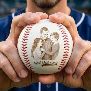 Könnte beinhalten: Ein Baseball mit einem Familienporträt und dem Text "Best Dad Ever". Der Ball hat rote Nähte und eine cremefarbene Oberfläche. Das Bild zeigt eine Person, die den Ball in ihren Händen hält.