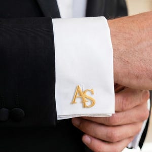 May include: A black suit jacket with a crisp white French cuff shirt sleeve. The cuff is fastened with a gold-toned metal cufflink featuring the stylized letters AS in a serif font, positioned prominently on the white fabric of the shirt cuff.