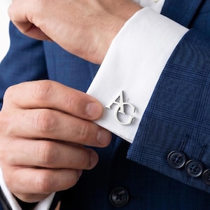 May include: A pair of silver-toned metal cufflinks featuring the letters AG in a stylized, connected font. The cufflinks are attached to the white cuffs of a long-sleeved shirt worn under a dark blue plaid blazer with three buttons on the sleeve.