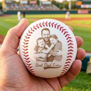 Può includere: Una palla da baseball bianca con cuciture rosse e un ritratto di famiglia inciso. La palla da baseball ha la scritta "Best Dad Ever" ed è tenuta in mano, con un campo da baseball sullo sfondo.