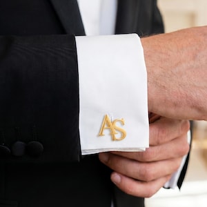 Puede incluir: Una chaqueta de traje negra con una manga de camisa blanca de puño doble. El puño está sujeto con un gemelo de metal dorado que presenta las letras estilizadas AS en una fuente con serifa, posicionado sobre la tela blanca de la camisa.