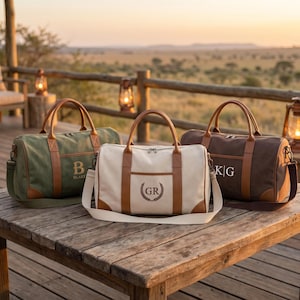 May include: Three canvas duffel bags with brown leather-like trim and handles. The olive green bag features the letter B and the name Blake. The cream bag displays the initials GR in a laurel wreath. The dark brown bag shows the initials K|G.