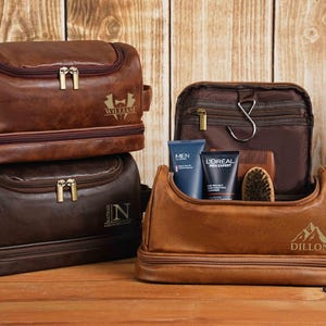 May include: Leather toiletry bags in shades of brown, stacked on a wooden surface. One bag is open, displaying grooming products. The bags have gold zippers and personalized text. The open bag contains blue tubes, a brush, and a brown sponge.