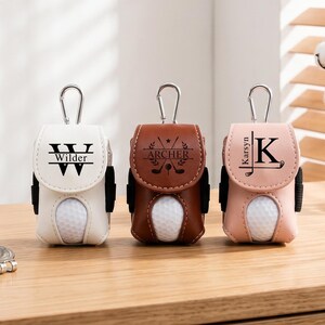 May include: Three golf ball pouches in white, brown, and light pink, each featuring a silver carabiner, a cutout for a golf ball, and personalized black text. The white pouch says Wilder, the brown pouch says Archer, and the pink pouch says Karsyn.