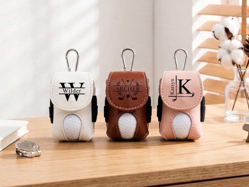 May include: Three golf ball pouches in white, brown, and light pink, each featuring a silver carabiner, a cutout for a golf ball, and personalized black text. The white pouch says Wilder, the brown pouch says Archer, and the pink pouch says Karsyn.