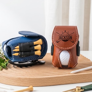 May include: A blue golf ball pouch holding three yellow tees and a brown golf ball pouch featuring the name JACKSON engraved on the flap with a golf ball visible inside. Both pouches have white stitching and a black clip on the side.