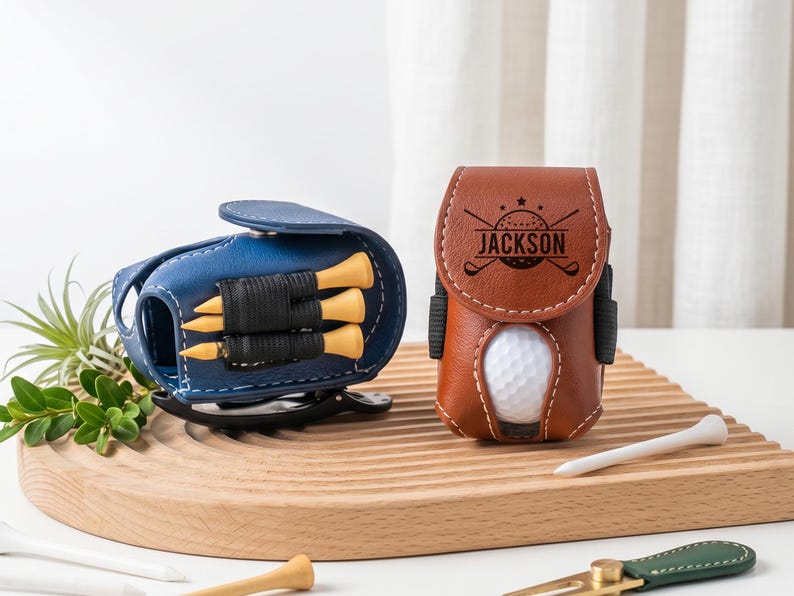 May include: A blue golf ball pouch holding three yellow tees and a brown golf ball pouch featuring the name JACKSON engraved on the flap with a golf ball visible inside. Both pouches have white stitching and a black clip on the side.