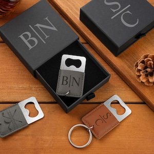 Könnte beinhalten: Eine Sammlung personalisierter Flaschenöffner und Schlüsselanhänger. Ein Flaschenöffner befindet sich in einer schwarzen Box mit den Initialen "BN". Zwei Schlüsselanhänger haben die Initialen "BN" und "SC". Ein Schlüsselanhänger hat Golfschläger-Grafiken. Die Artikel befinden sich auf einer Holzoberfläche.