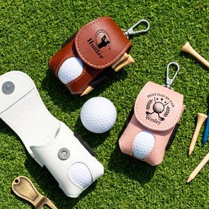 May include: Three golf ball pouches in brown, pink, and white leather-like material with metal clips. The brown pouch features the name Hunter, the pink pouch says Best Dad By Par Wesley, and all contain white golf balls and wooden golf tees.