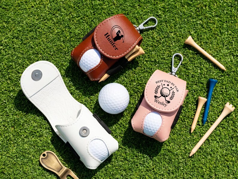 May include: Three golf ball pouches in brown, pink, and white leather-like material with metal clips. The brown pouch features the name Hunter, the pink pouch says Best Dad By Par Wesley, and all contain white golf balls and wooden golf tees.