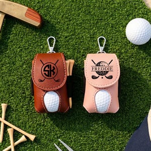 May include: Two golf ball pouches with silver metal clips, one brown and one light pink. The brown pouch features a black circular logo with the letters SK and crossed golf clubs. The pink pouch displays a golf ball graphic with the name FREDDIE.