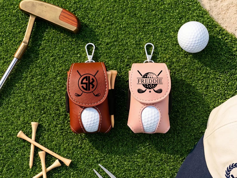 May include: Two golf ball pouches with silver metal clips, one brown and one light pink. The brown pouch features a black circular logo with the letters SK and crossed golf clubs. The pink pouch displays a golf ball graphic with the name FREDDIE.