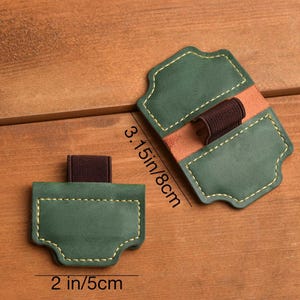 May include: Two green leather pen holders with brown elastic bands and tan leather accents. The pen holders are 2 inches (5 cm) wide and 3.15 inches (8 cm) long. The edges are stitched with yellow thread.